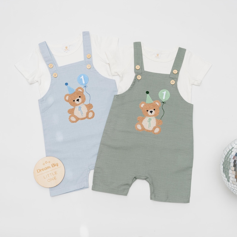 May include: Two short-sleeved rompers with overalls, one in light blue and one in sage green, each layered over a white t-shirt. Both feature an embroidered brown teddy bear wearing a party hat and holding a balloon with the number 1.