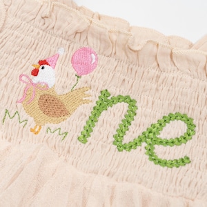 May include: A light beige fabric with an embroidered design. The design features a cartoon chicken wearing a party hat and holding a pink balloon, with the word "one" in green. The fabric has a ruffled edge.