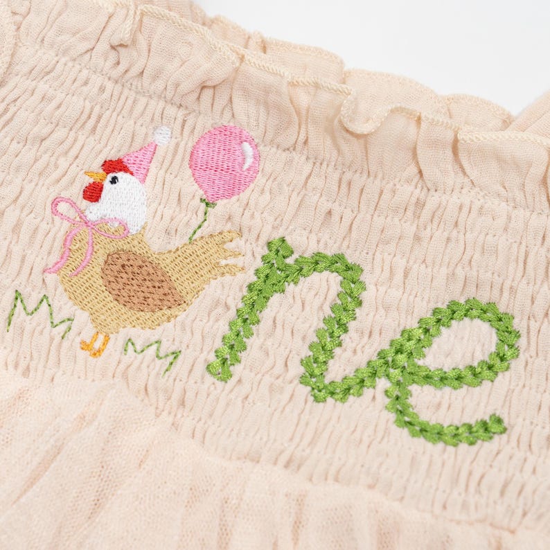 May include: A light beige fabric with an embroidered design. The design features a cartoon chicken wearing a party hat and holding a pink balloon, with the word "one" in green. The fabric has a ruffled edge.