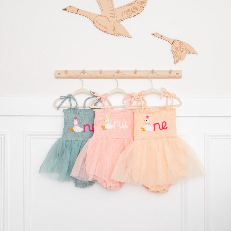 May include: Three baby dresses in pastel colors: teal, pink, and peach. Each dress has a tulle skirt and a bodice embroidered with a duck and the word "one". The dresses hang on a wooden rack, with wooden geese decorations above.