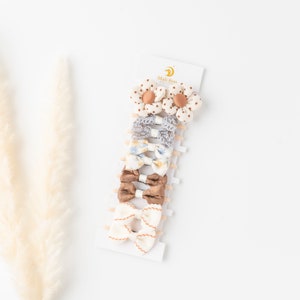May include: Set of 7 hair clips with bows and flowers. The clips are in shades of brown, white, and blue. The flowers are white with brown polka dots. The bows are made of fabric with floral patterns. The clips are displayed on a white card with the text "Mali Boo" in gold.