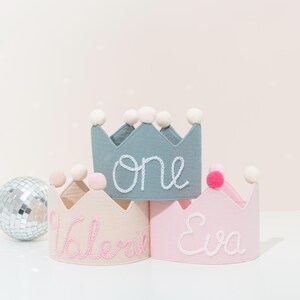 May include: Three fabric crowns in pastel colors: light blue, beige, and pink. Each crown has a different name embroidered on it in a contrasting color. The light blue crown says "one". Each crown is topped with white pom-poms.