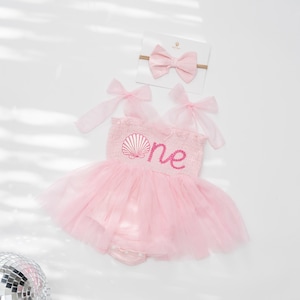 May include: A pink baby romper with a tulle skirt, featuring a seashell design and the word "one" embroidered in pink. A matching pink bow headband is included. The outfit is designed for a baby's first birthday.
