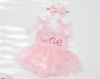 May include: A pink baby romper with a tulle skirt, featuring a seashell design and the word "one" embroidered in pink. A matching pink bow headband is included. The outfit is designed for a baby's first birthday.