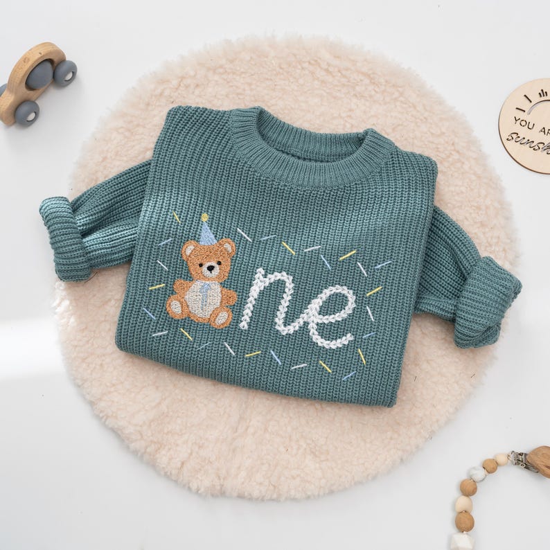 May include: A teal knitted sweater with long sleeves. The sweater features an embroidered teddy bear wearing a party hat and the word "one" in white script. Colorful confetti-like accents surround the text and bear.
