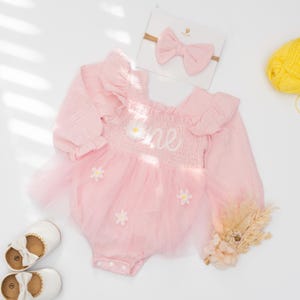 以下が含まれることがあります： ピンク色のベビー服。長袖、チュールスカート、デイジーのアクセント付き。「one」の文字がボディスに刺繍されています。お揃いのリボンヘアバンドと白いベビーシューズ（リボン付き）が含まれています。ドライフラワーと黄色の毛糸玉も写っています。