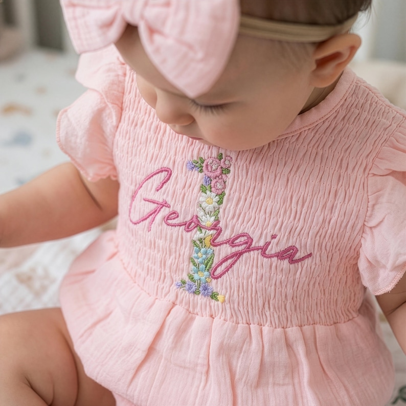 K&ouml;nnte beinhalten: Ein hellrosa Baby-Strampler mit R&uuml;schen&auml;rmeln und gesmoktem Oberteil. In der Mitte befindet sich eine gestickte florale Eins mit dem Namen Georgia in dunkelrosa Schreibschrift. Ein passendes rosa Stirnband mit Schleife wird auf dem Kopf getragen.