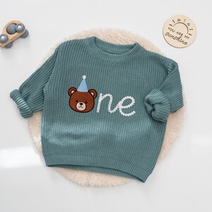 K&ouml;nnte beinhalten: T&uuml;rkisfarbener Strickpullover mit einem braunen B&auml;ren mit blauem Partyhut, der auf der Vorderseite gestickt ist. Das Wort "one" ist in wei&szlig;er Schrift darunter gestickt. Der Pullover hat lange &Auml;rmel und einen Rundhalsausschnitt.