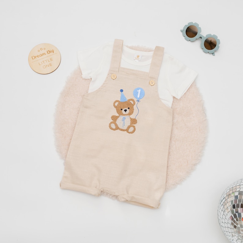 May include: A beige overall short set with a white t-shirt. The overalls feature an embroidered brown teddy bear wearing a blue party hat and holding a blue balloon with the number 1. A wooden disc reads Dream Big Little One.