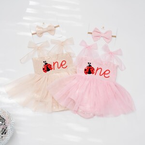 May include: Two baby girl outfits, one in light pink and the other in beige, each with a tutu skirt and a headband. The outfits feature a ladybug design and the word "one" embroidered on the bodice. The headbands have a matching bow.