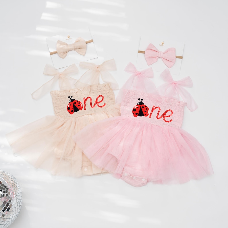 May include: Two baby girl outfits, one in light pink and the other in beige, each with a tutu skirt and a headband. The outfits feature a ladybug design and the word "one" embroidered on the bodice. The headbands have a matching bow.