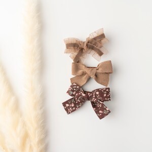 May include: Three brown hair bows, one with a floral pattern, one with a corduroy texture, and one with a sheer fabric. The bows are arranged vertically on a white background.