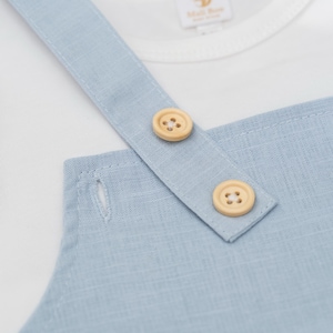 May include: A light blue overall garment featuring a textured fabric, two round light-colored wooden buttons on the shoulder strap, and a visible buttonhole. A white shirt is layered underneath with a tag reading Mali Boo.
