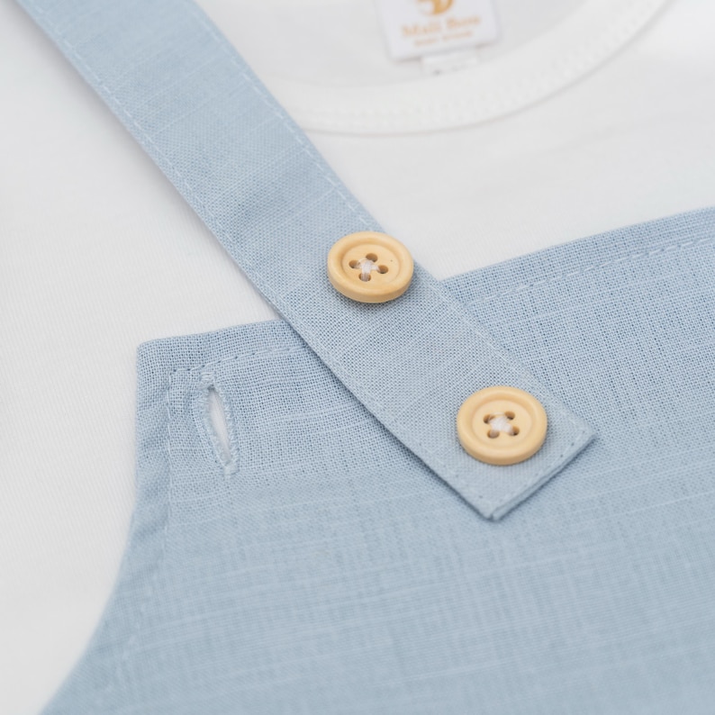 May include: A light blue overall garment featuring a textured fabric, two round light-colored wooden buttons on the shoulder strap, and a visible buttonhole. A white shirt is layered underneath with a tag reading Mali Boo.
