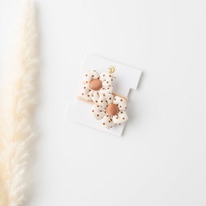 May include: Two white fabric hair clips with brown polka dots and a brown button center. The clips are on a white card with the word "Boo" printed on it.