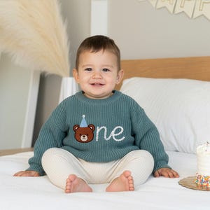 K&ouml;nnte beinhalten: Ein tealfarbener Strickpullover mit einem braunen B&auml;rengrafik, der eine blaue Partyhut tr&auml;gt und das Wort "one" in Wei&szlig; gestickt hat. Der Pullover wird mit cremefarbenen Hosen kombiniert. Eine Geburtstagstorte mit Streuseln ist sichtbar.
