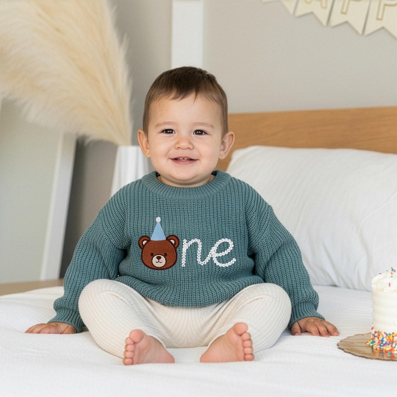 K&ouml;nnte beinhalten: Ein tealfarbener Strickpullover mit einem braunen B&auml;rengrafik, der eine blaue Partyhut tr&auml;gt und das Wort "one" in Wei&szlig; gestickt hat. Der Pullover wird mit cremefarbenen Hosen kombiniert. Eine Geburtstagstorte mit Streuseln ist sichtbar.