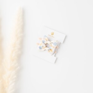 May include: Two small floral hair bows on a white card with the text "Mali Boo" in gold lettering. The bows are made of a white fabric with a blue and yellow floral pattern.