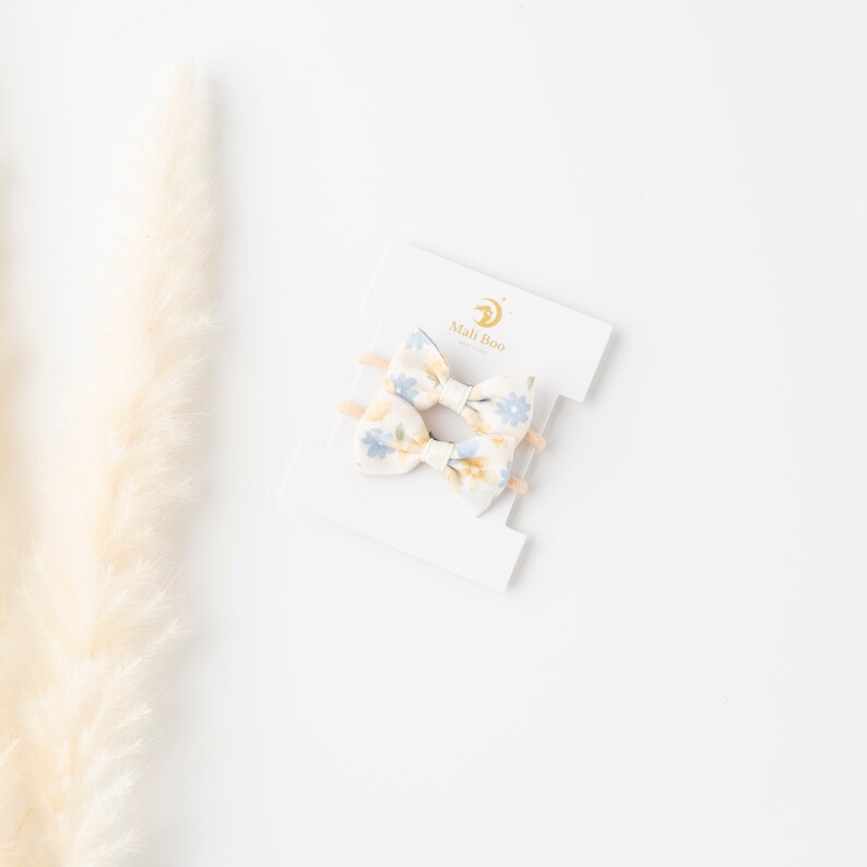 May include: Two small floral hair bows on a white card with the text "Mali Boo" in gold lettering. The bows are made of a white fabric with a blue and yellow floral pattern.