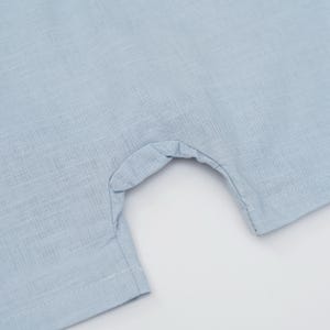 May include: A light blue garment featuring a woven texture and a curved cutout section. The fabric displays a visible cross-hatch pattern with straight stitched seams along the edges of the opening and the hemline of the textile material.