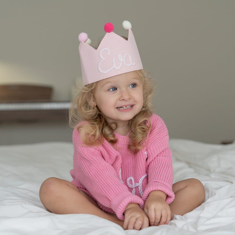 May include: A pink felt crown with the name "Eva" embroidered in white, adorned with pink and white pom-poms. The crown is worn by a child wearing a pink sweater. The child is sitting on a white bed.