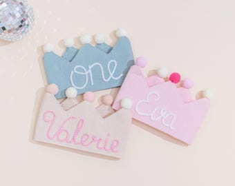 May include: Three fabric crowns in pastel colors: light blue, pink, and pale pink. Each crown has a scalloped edge and is adorned with small pom-poms. The crowns are embroidered with the names "one", "Eva", and "Valerie" in white and pink thread.
