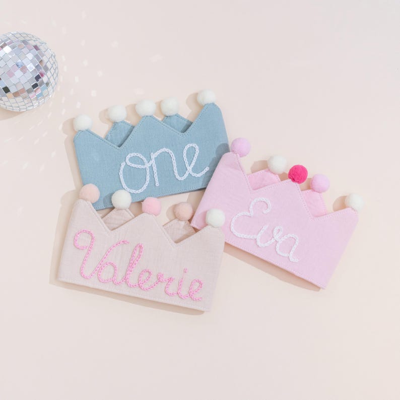 May include: Three fabric crowns in pastel colors: light blue, pink, and pale pink. Each crown has a scalloped edge and is adorned with small pom-poms. The crowns are embroidered with the names "one", "Eva", and "Valerie" in white and pink thread.