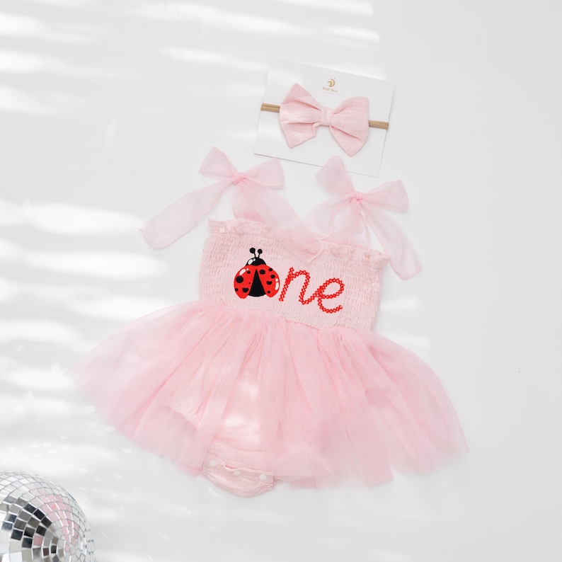 May include: Pink baby romper with a tulle skirt and a matching headband. The romper features a ladybug design and the word "one" in red. The headband has a pink bow. Perfect for a first birthday.