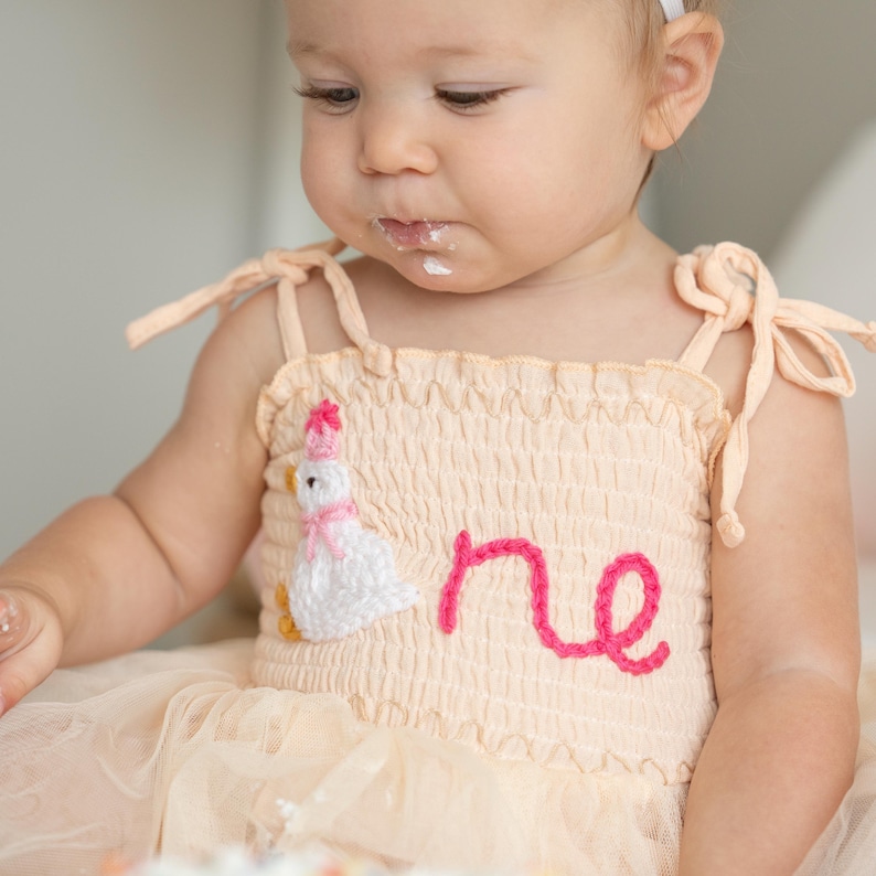 May include: A light peach-colored dress with a white crochet duck and the word "one" in pink. The dress has tie straps and a tulle skirt. The child is wearing a white headband and has frosting on their face.