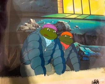 May include: Two Teenage Mutant Ninja Turtles wearing winter hats and puffy jackets are crouched down in a snowy scene. The turtle on the left is wearing a purple hat and the turtle on the right is wearing an orange hat.