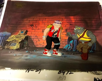 May include: A hand-painted animation cel featuring two characters from the Teenage Mutant Ninja Turtles cartoon. The characters are a pig-like creature wearing a red vest and a rhino-like creature wearing a yellow vest. The cel is set against a brick wall background.