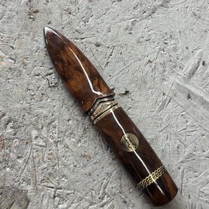 May include: A fountain pen with a polished dark brown wood grain body and a matching cap. The pen features ornate brass-colored metal accents, including a decorative band and a circular inlay on the barrel with the text Beyoglu engraved in script.