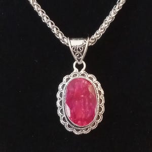 May include: A silver necklace with a large, oval, ruby-colored pendant. The pendant is set in a decorative silver frame. The chain is a woven design. The necklace is displayed on a black velvet background.