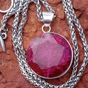 May include: A silver necklace with a large, faceted, round, deep pink pendant. The pendant is set in a silver bezel and hangs from a thick, woven silver chain. The necklace has a toggle clasp.