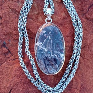 May include: A silver necklace with an oval pendant. The pendant features a polished stone with a mix of dark and light colors. The chain is a woven design with a toggle clasp. The necklace is displayed on a reddish-brown surface.