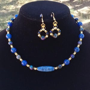 May include: A necklace and matching pair of drop earrings featuring round blue and dark mottled beads, small gold-toned spacer beads, and a central elongated blue cylindrical bead. The jewelry set is displayed on a black stand.