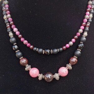 May include: A double-strand necklace featuring a mix of matte black beads, faceted magenta beads, and smooth pink and dark brown spherical beads. The strands are accented with small silver-toned metal spacers and textured silver-toned bead caps.