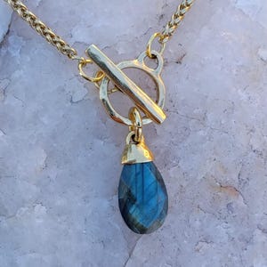 May include: Gold-toned necklace featuring a teardrop-shaped blue gemstone pendant. The pendant is suspended from a gold toggle clasp and a delicate chain. The gemstone has a faceted surface, catching the light.