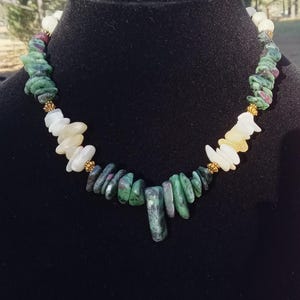 May include: A gemstone necklace featuring a variety of natural stones in shades of green, white, yellow, and red. The necklace is displayed on a black mannequin and has gold-colored accents.