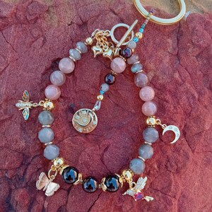 May include: A beaded keychain with a gold ring. The keychain features a variety of beads in shades of pink, gray, and black, with gold accents. Charms include a dragonfly, butterfly, hummingbird, moon, and stars.
