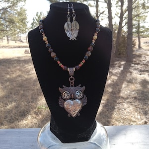 May include: A steampunk-style necklace and earring set. The necklace features a pendant shaped like an owl with a heart and gear details. The beaded necklace includes brown, black, and red beads. The earrings have a wing design. The jewelry is displayed on a black bust.