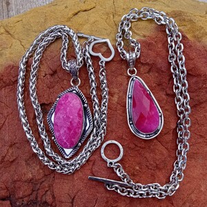 May include: Two silver necklaces with large, vibrant pink gemstone pendants. One pendant is a teardrop shape, the other is an elongated oval. The chains are thick and textured, with a toggle clasp.