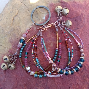 May include: Multi-strand beaded bracelet with a key ring and multiple bells. The bracelet features a variety of colors, including red, green, and gold. The beads are made of glass and metal.
