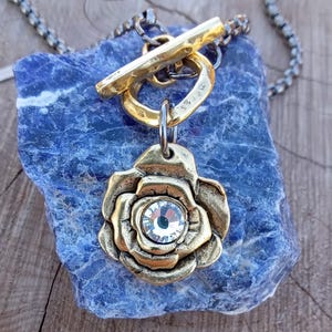 May include: A gold-toned pendant necklace featuring a rose design with a clear crystal center. The necklace has a toggle clasp and a silver-toned chain. The pendant is displayed on a blue stone surface, creating a contrast of colors and textures.