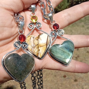 May include: Three heart-shaped pendant necklaces with silver chains. Each pendant features a different gemstone: a green-grey stone, a cream and brown stone, and a blue-green stone. Each pendant has a silver frame and is adorned with a small gemstone.