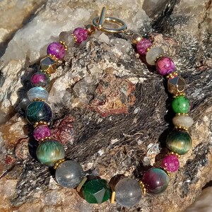 May include: A beaded bracelet featuring a variety of colorful gemstones. The bracelet includes round, faceted, and geometric beads in shades of green, purple, gold, and gray. The clasp is gold-toned.