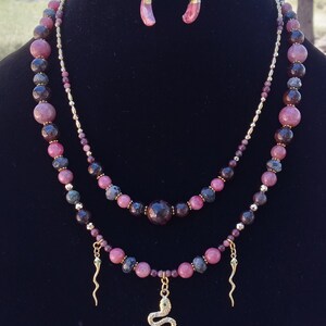 May include: A layered beaded necklace featuring pink and dark purple stones, gold-tone spacers, and three gold-tone snake charms. The set includes matching drop earrings with pink stone accents, all displayed on a black velvet bust form.