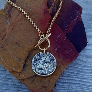 May include: A gold-toned necklace with a circular pendant featuring a sleeping fox under a crescent moon and trees. The pendant is attached to a gold chain with a toggle clasp. The necklace is displayed on a textured, reddish-brown stone.