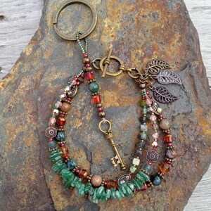 May include: A multi-strand beaded bracelet with a key charm and leaf accents. The bracelet features green, red, and brown beads, with a bronze-colored clasp and key. The key ring is attached to the top of the bracelet.