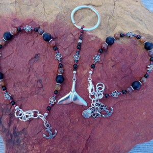 May include: A collection of beaded keychains and bracelets. The keychains feature a silver whale tail charm. The bracelets and keychains have black, brown, and red beads with silver flower accents. One keychain has a silver anchor charm.